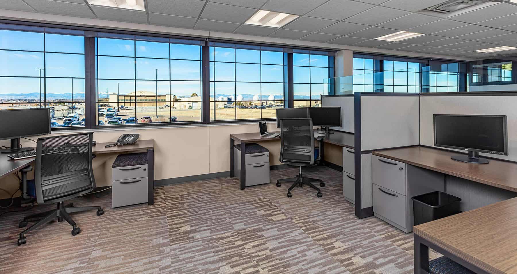 Office space with desks, cabinets, monitors, and chairs. Window shows a blue sky and the Colorado National Guard Air Force Base.
