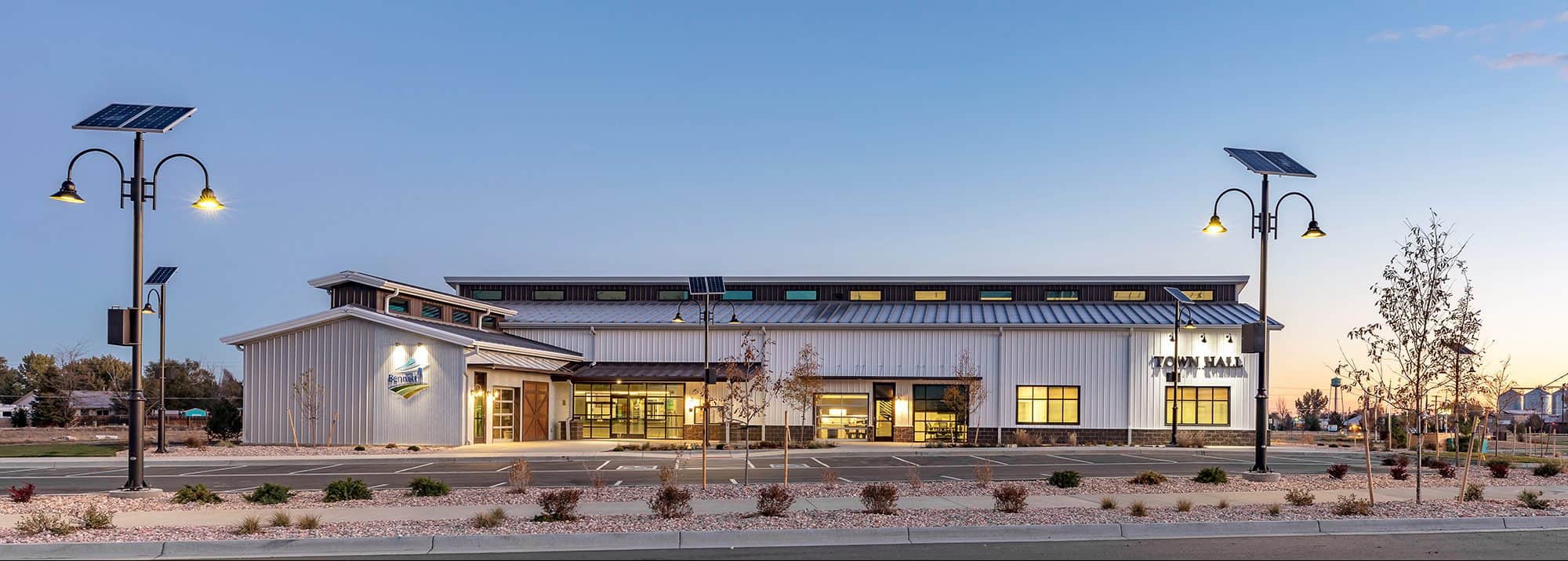 The entire exterior of the Town of Bennett Town Hall at dusk. The parking lot is well lit by lights.