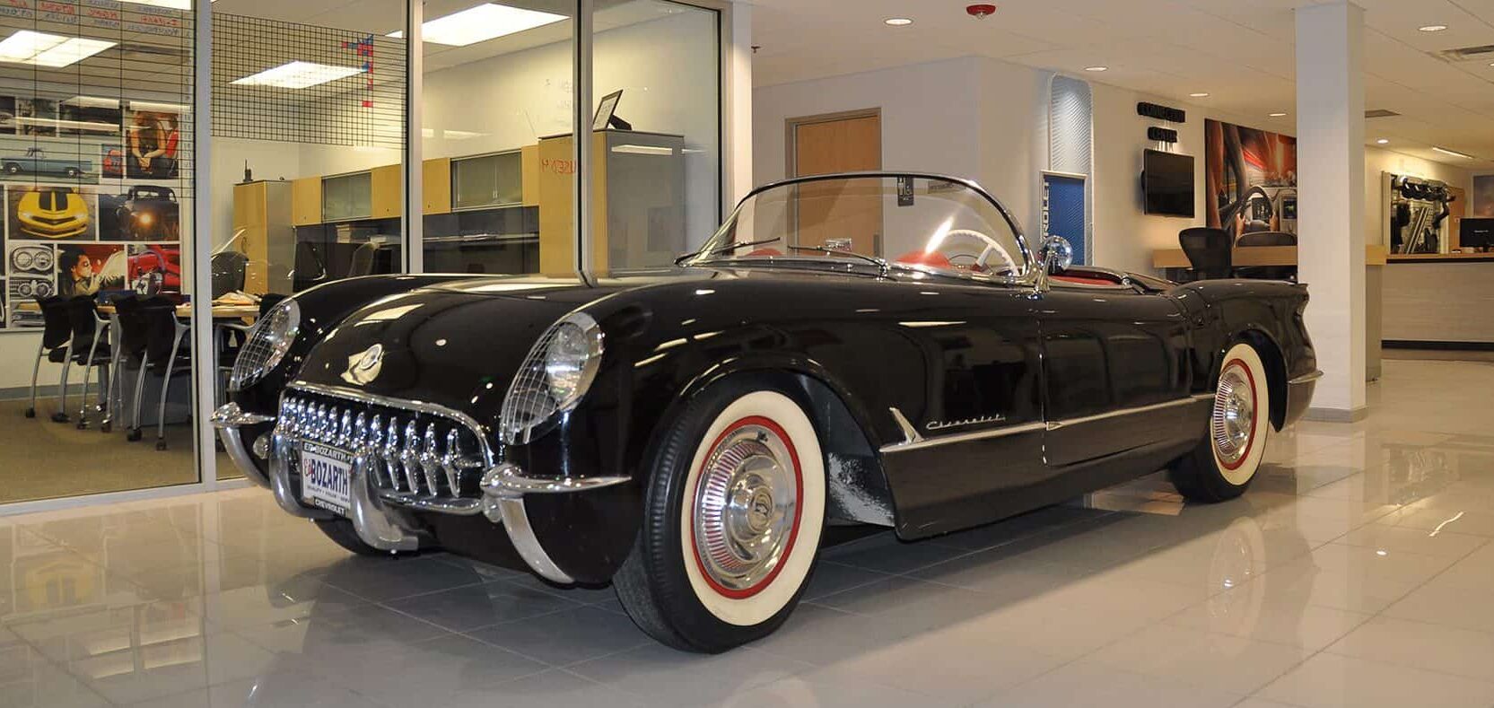 Reflective, classic sports car sitting on the floor of the office space. An office with floor to ceiling windows next to the car.