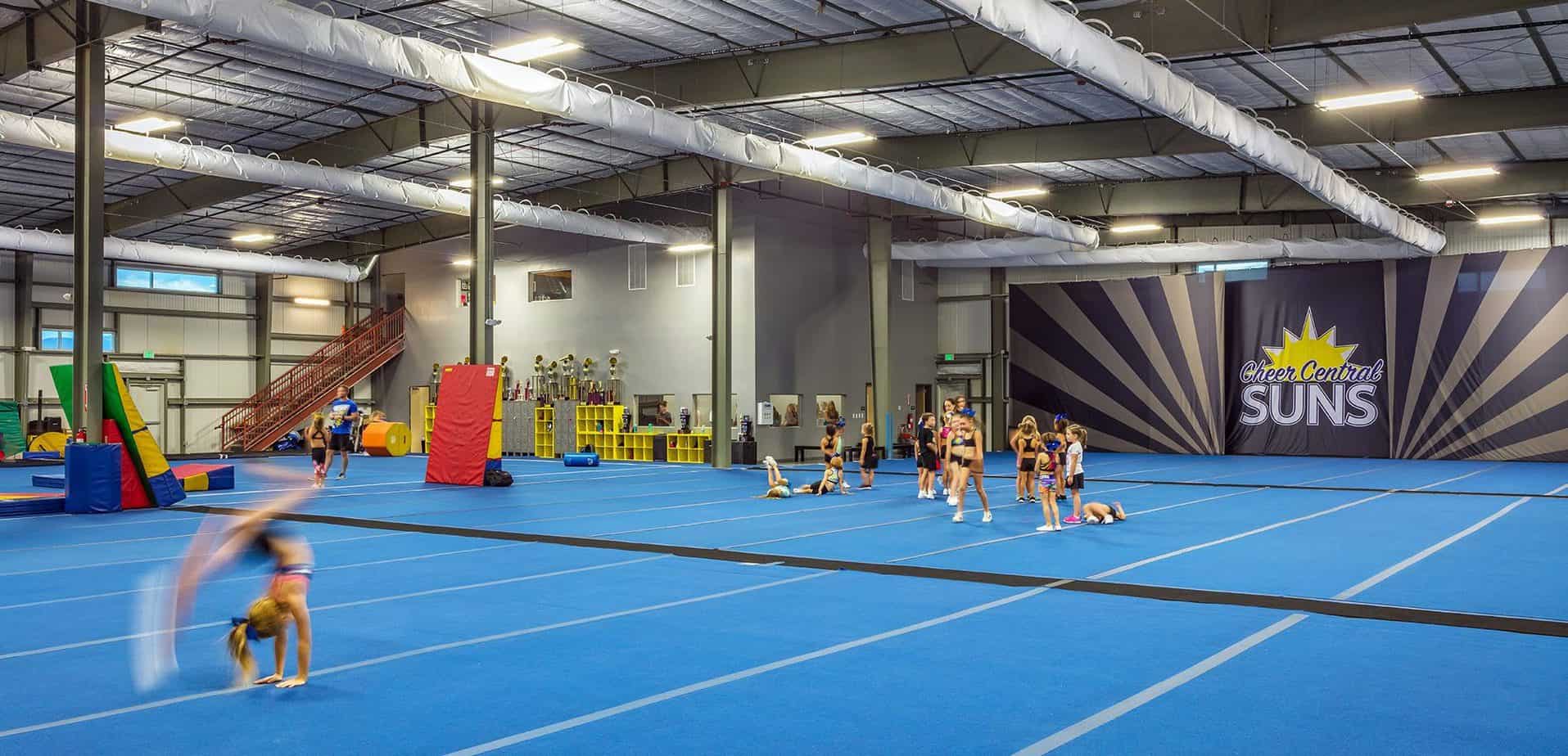 Large open, indoor practice area with blue mats. The "Suns" logo is on a large wall in the back and cheerleaders are practicing.
