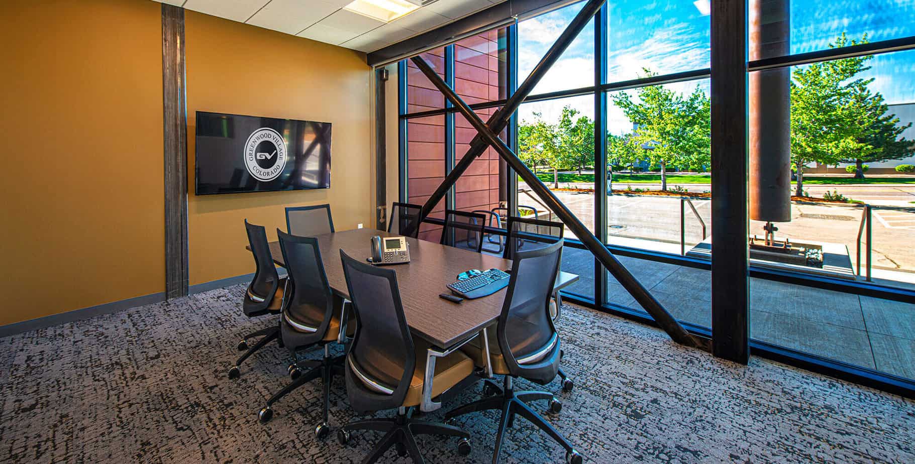 Conference room with rectangular table, chairs, large hanging monitor, and floor to ceiling windows that provide natural lighting.