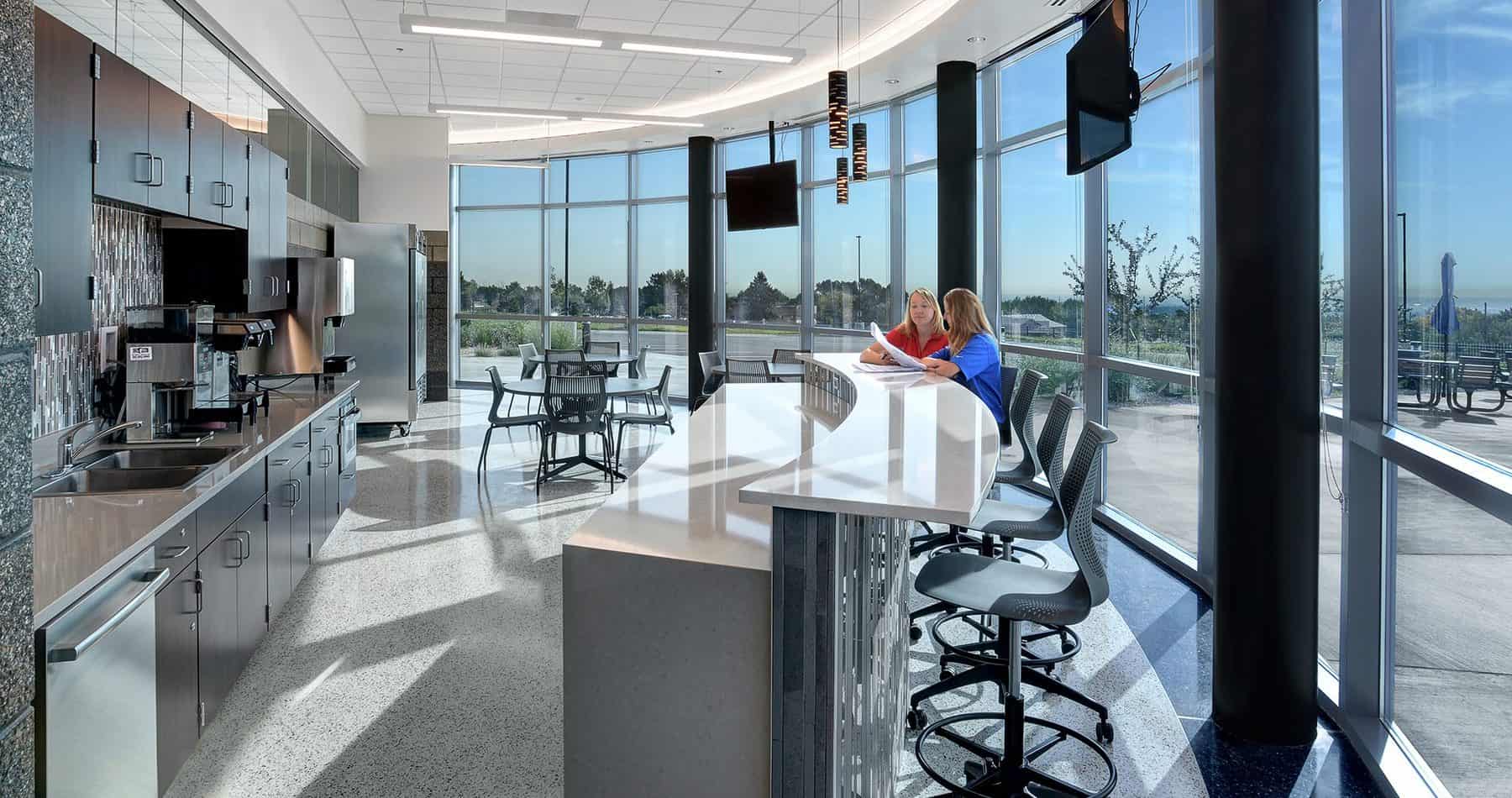 Naturally lit communal and kitchen space with a counter top and bar-top area with bar stools. There are TV monitors hanging and floor to ceiling windows.
