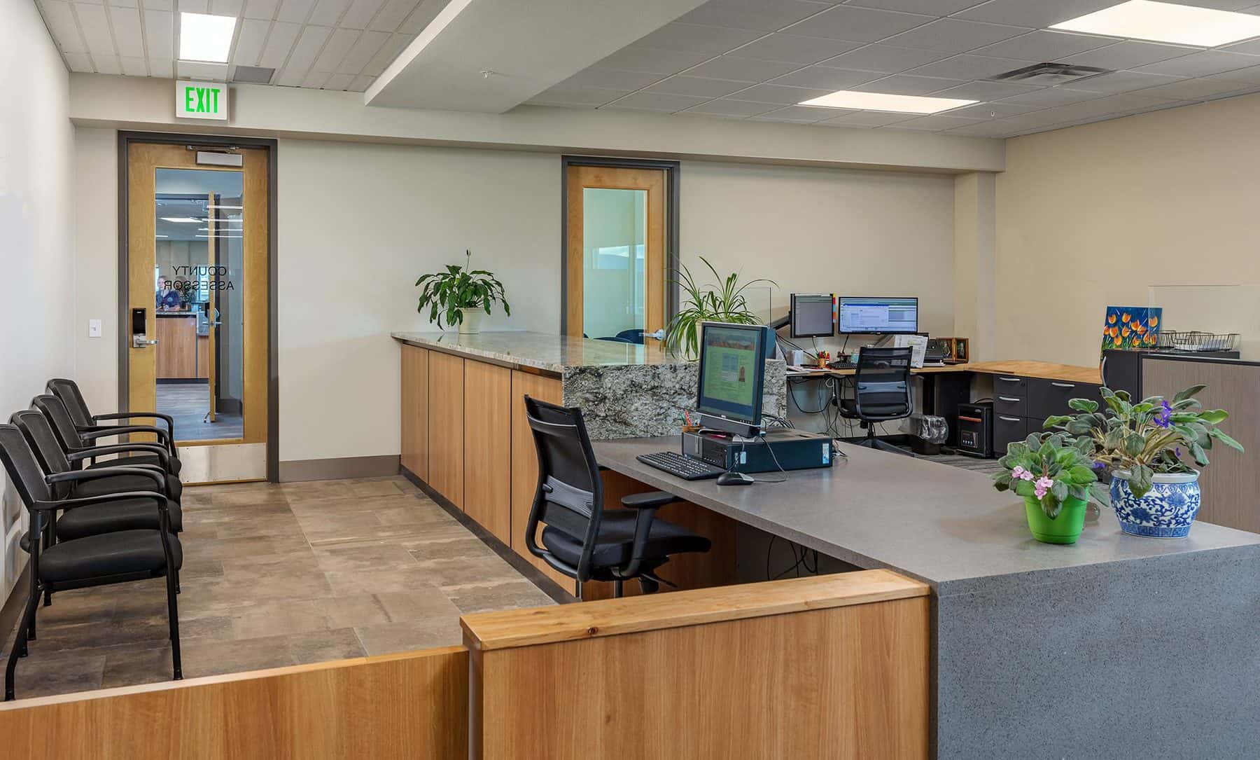 Office area with desks and chairs. A chair is in front of a lowered area of the reception desk. A computer monitor faces this chair