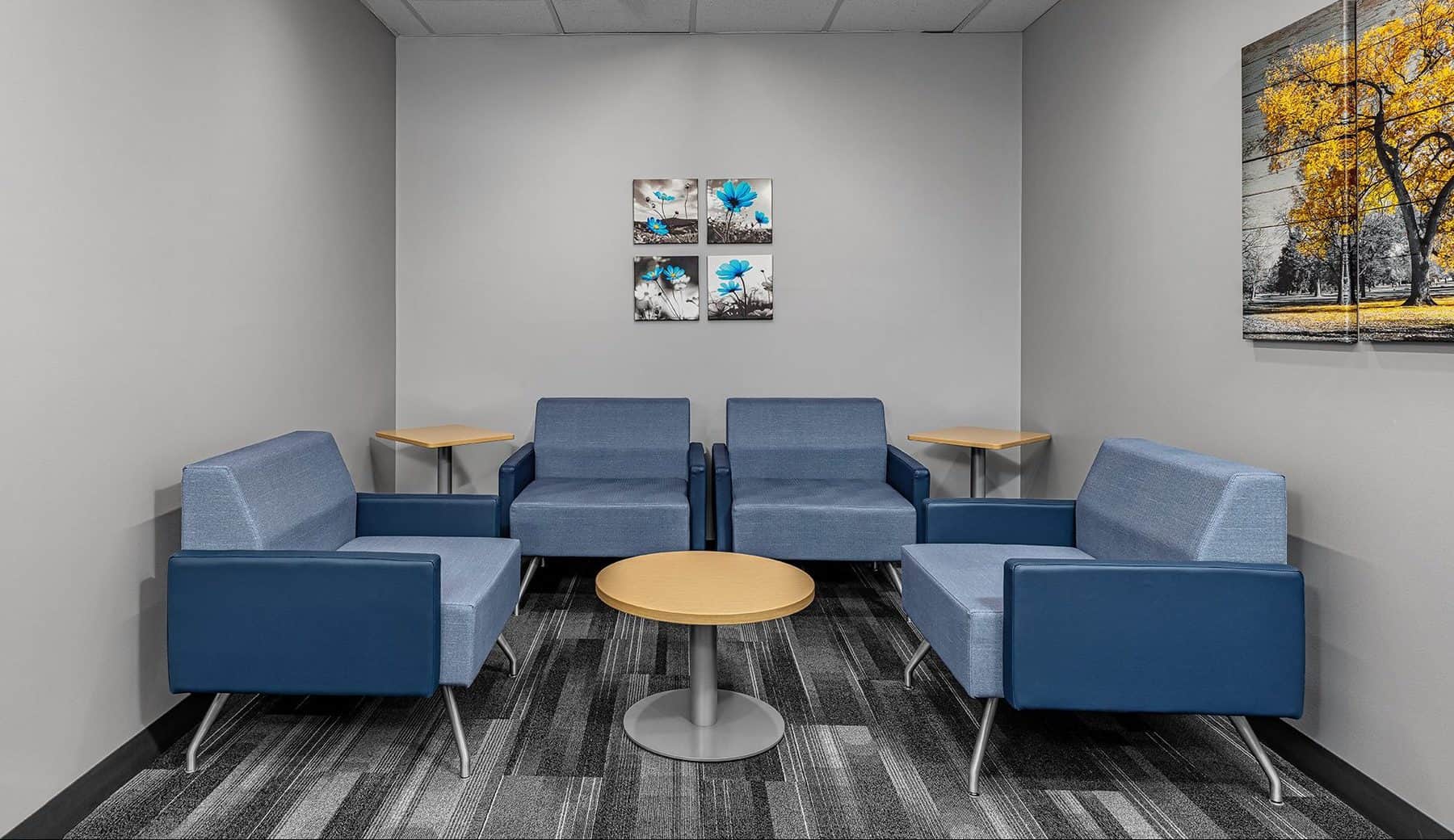 Waiting room for small groups with four comfy chairs around a circular table in the center of the room. Two paintings hang on the wall.