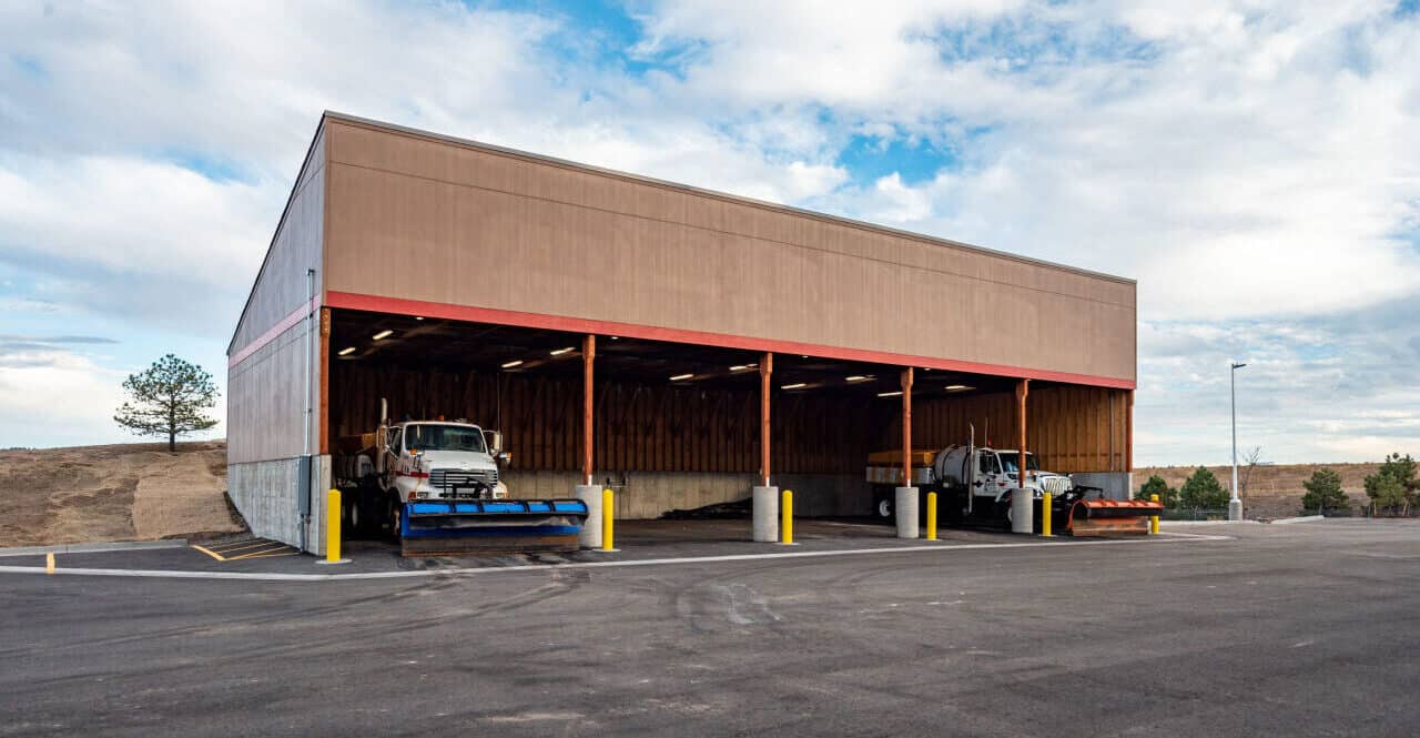 Exterior view of the large vehicle storage building with the bay door open and a 2 snow plow trucks inside.