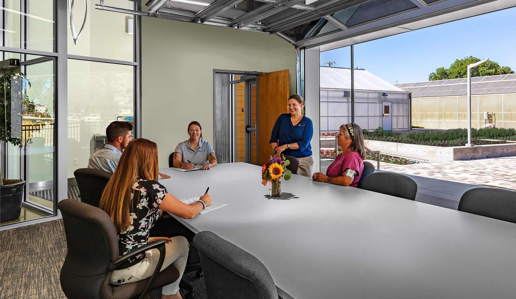 A group of City of Westminster greenhouse employees meet in a large conference room. The garage door to the conference room is open to the outside courtyard.